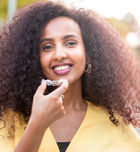 Woman with curly hair smiling while holding Invisalign aligner